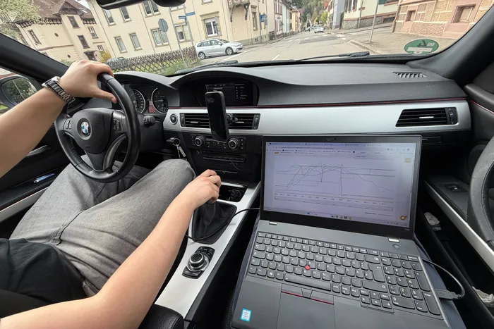 Blick vom Fahrersitz während der Kontrollfahrt: eine Hand am Lenkrad, auf dem Beifahrersitz ein Laptop mit mitlaufender Messkurve